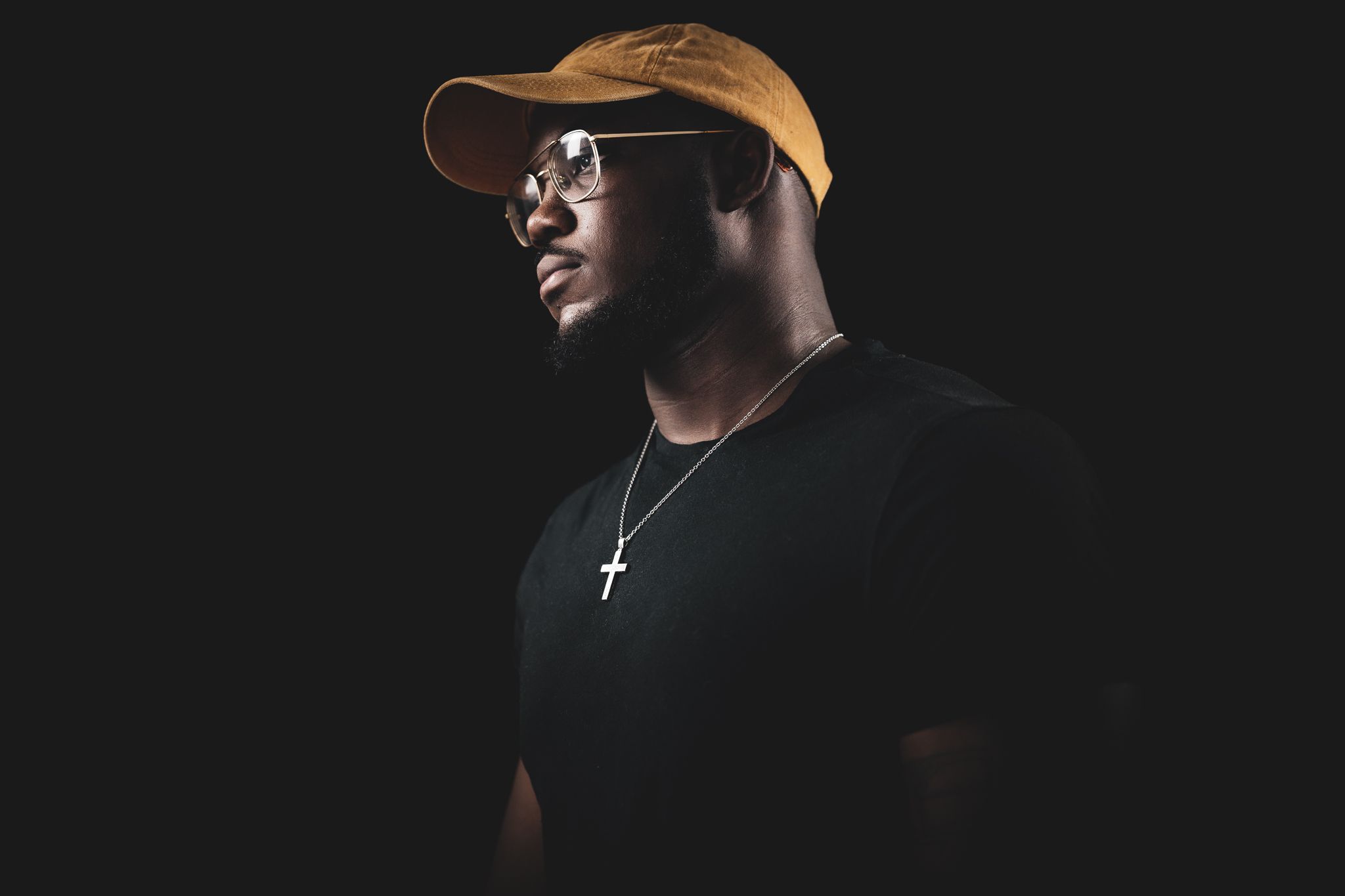A dramatic profile portrait of a person wearing sunglasses, baseball cap and cross necklace.