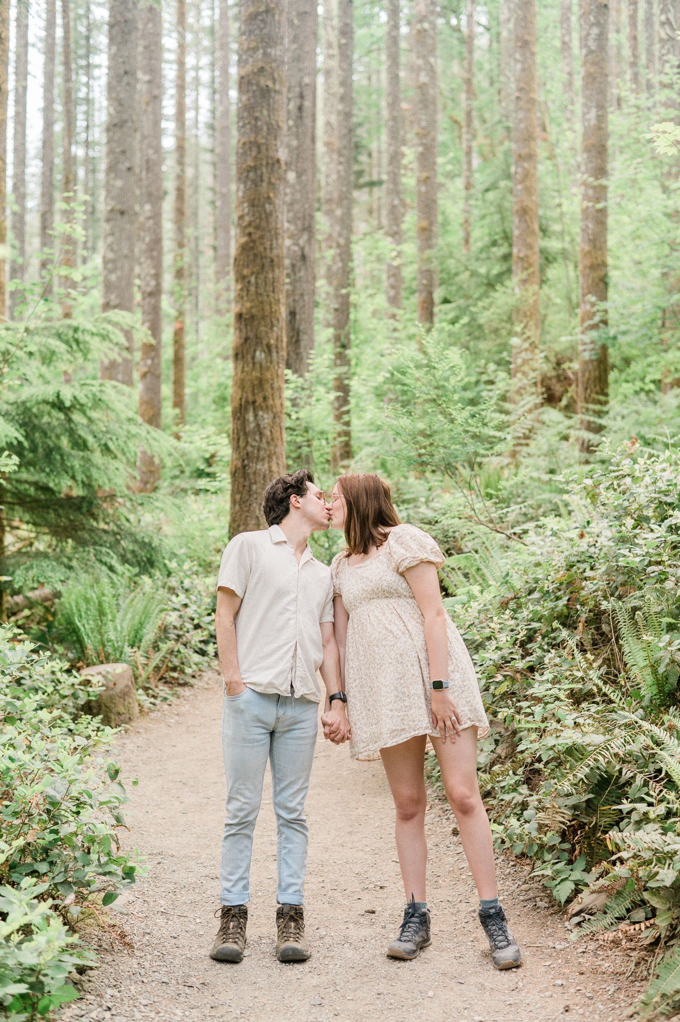 A couple kiss in peaceful forest trail surrounded by tall evergreen trees and lush ferns.