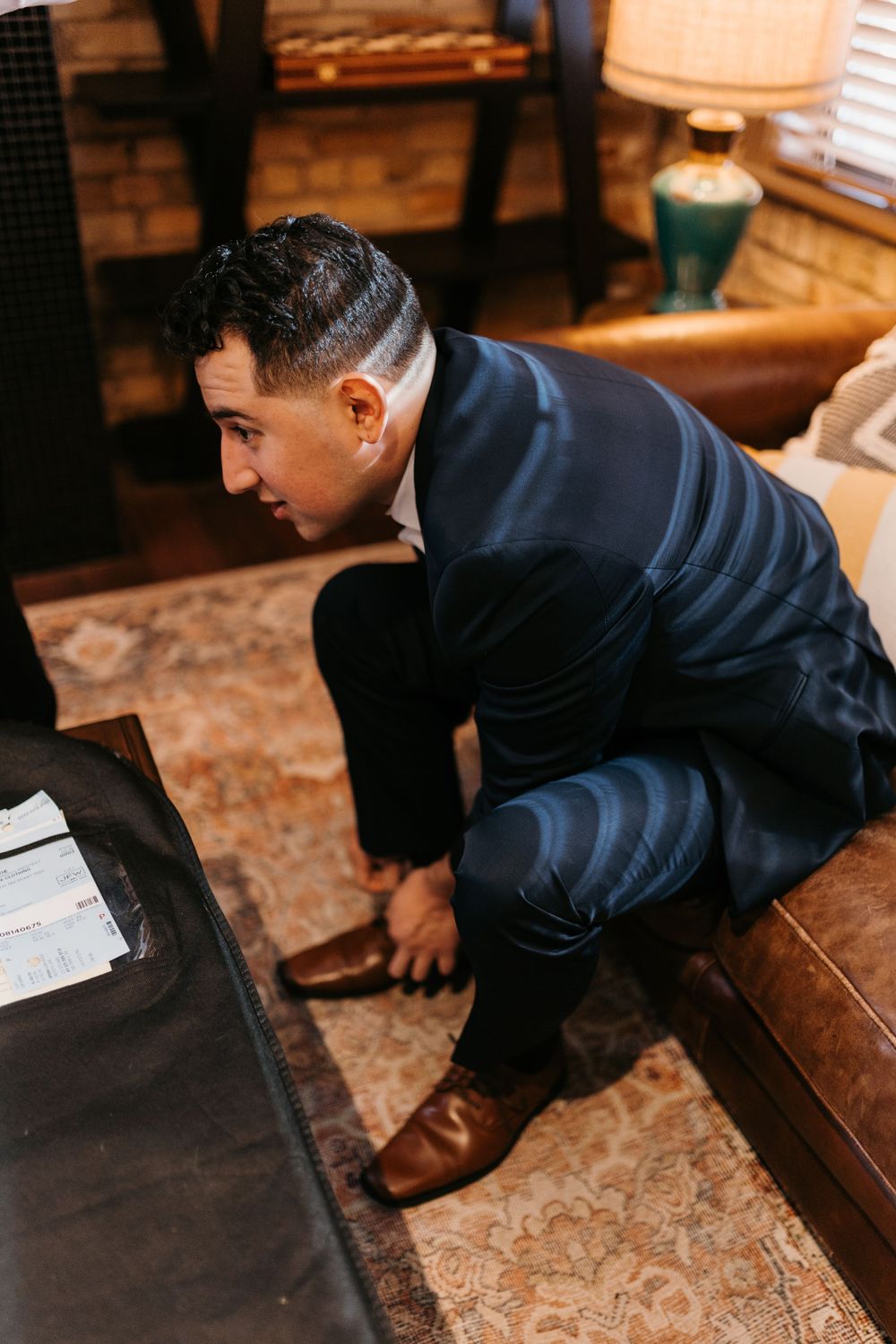 Groom in navy suit adjusting brown leather shoes while getting ready in warmly lit room.