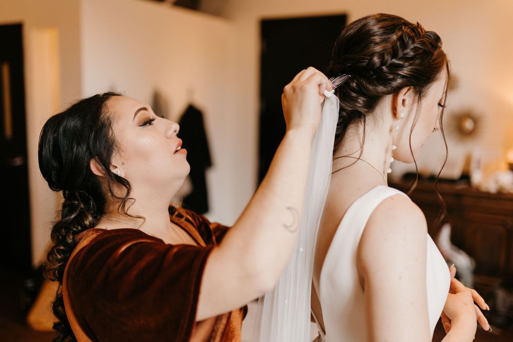 Bridesmaid in burgundy dress carefully attaching veil to bride's elegant updo hairstyle.