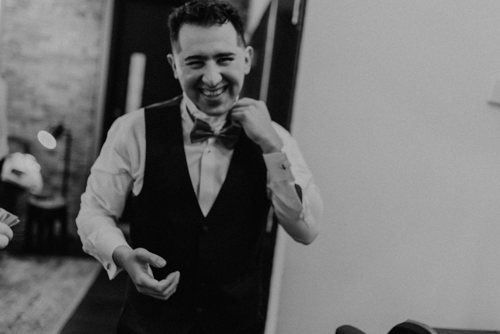 Smiling groom in black vest and bow tie laughing while getting dressed for his wedding day.