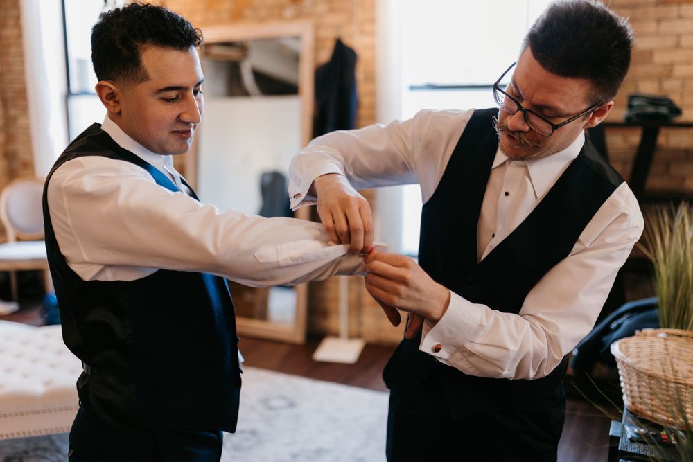 Two groomsmen in black vests help each other with cufflinks while getting ready for wedding.