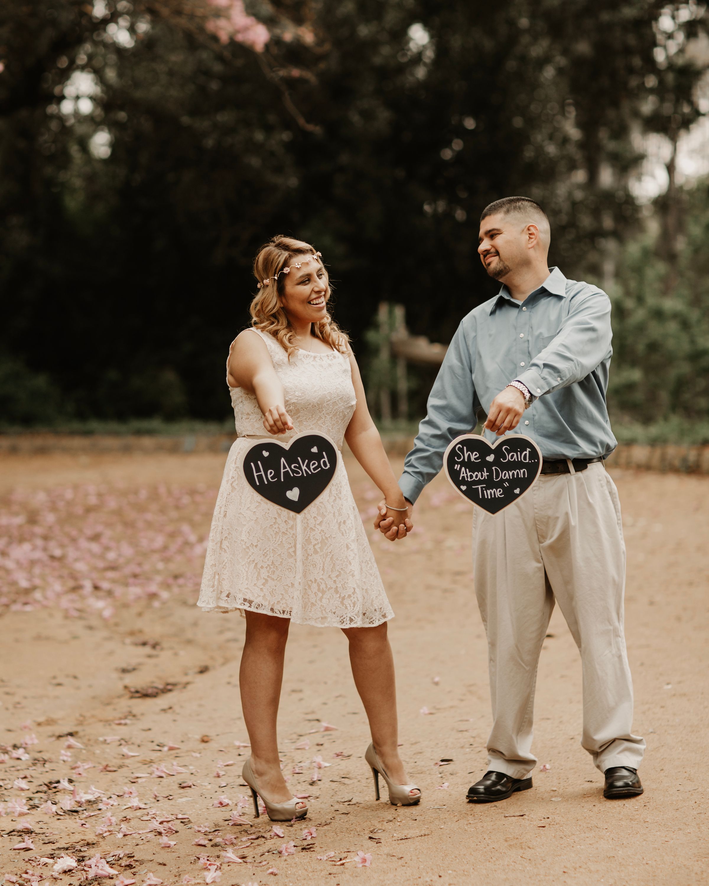 Couple holding heart-shaped chalkboard signs during an engagement photoshoot in a park.