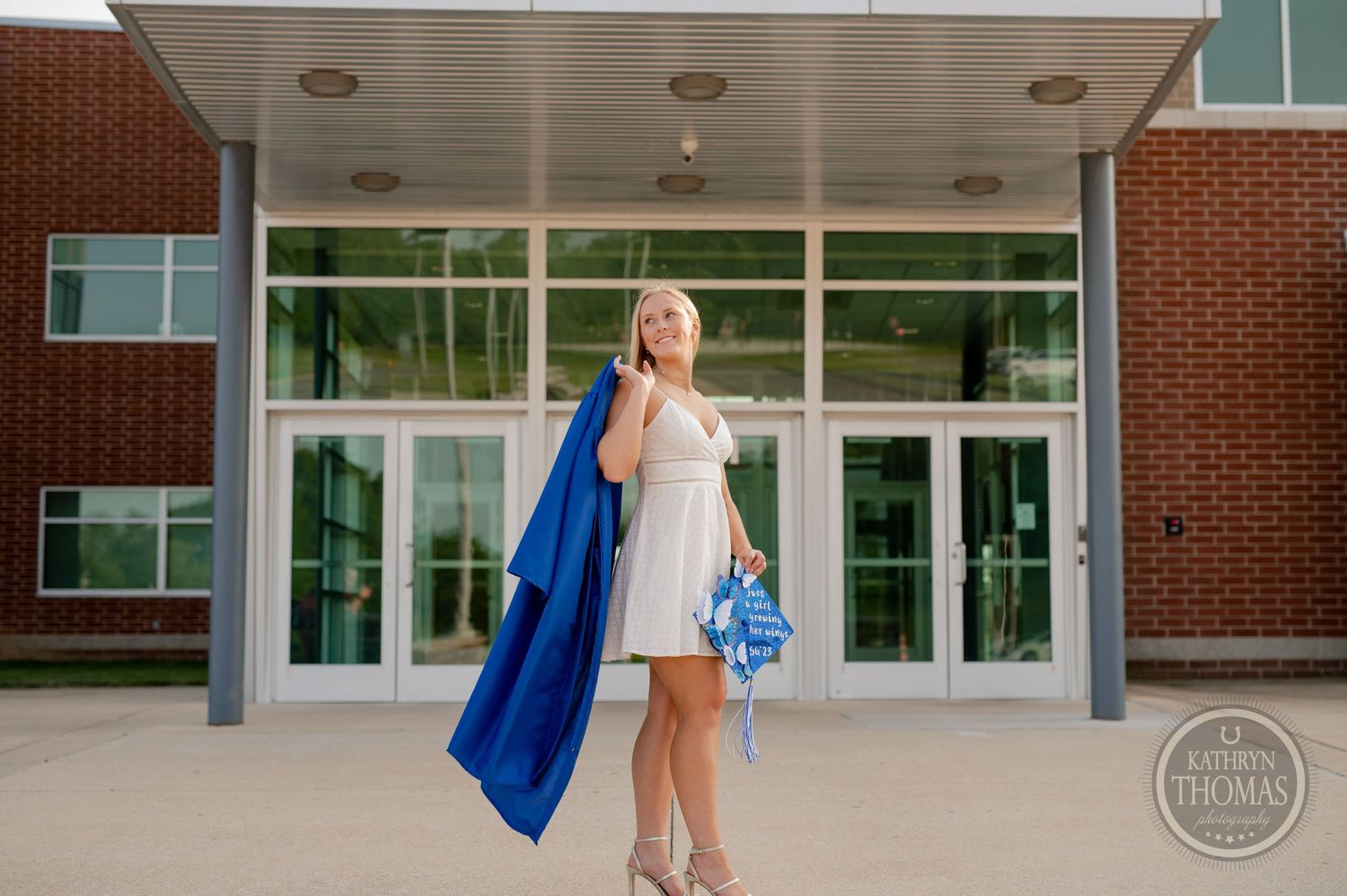 Graduate in blue gown and white dress poses at school entrance with modern glass doors.