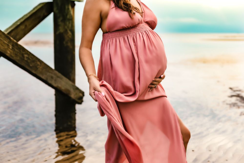 A flowing coral maternity dress detail against wooden dock posts on a sandy beach.