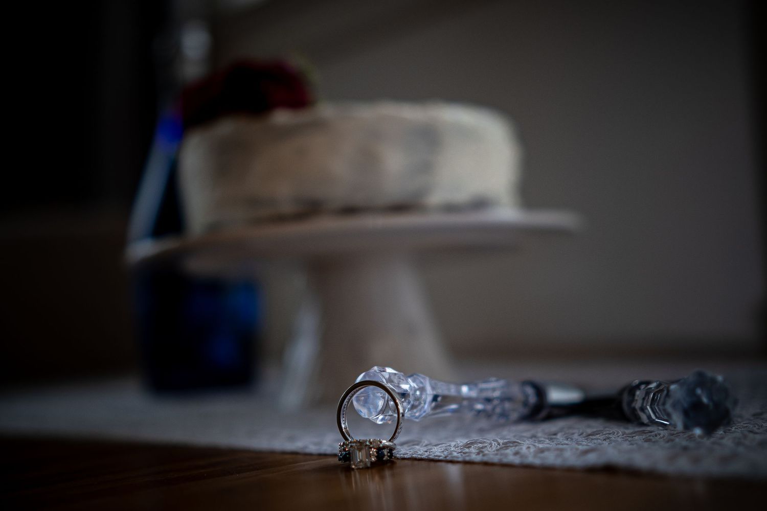 Wedding rings displayed on lace fabric with a white cake stand in soft focus background.