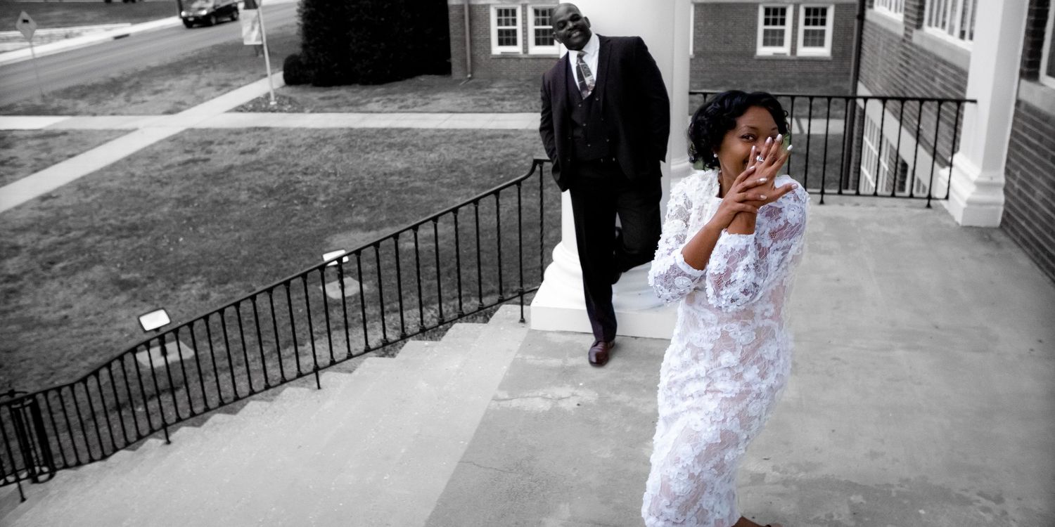 A person in white dress shows joyful emotion while another walks in background.