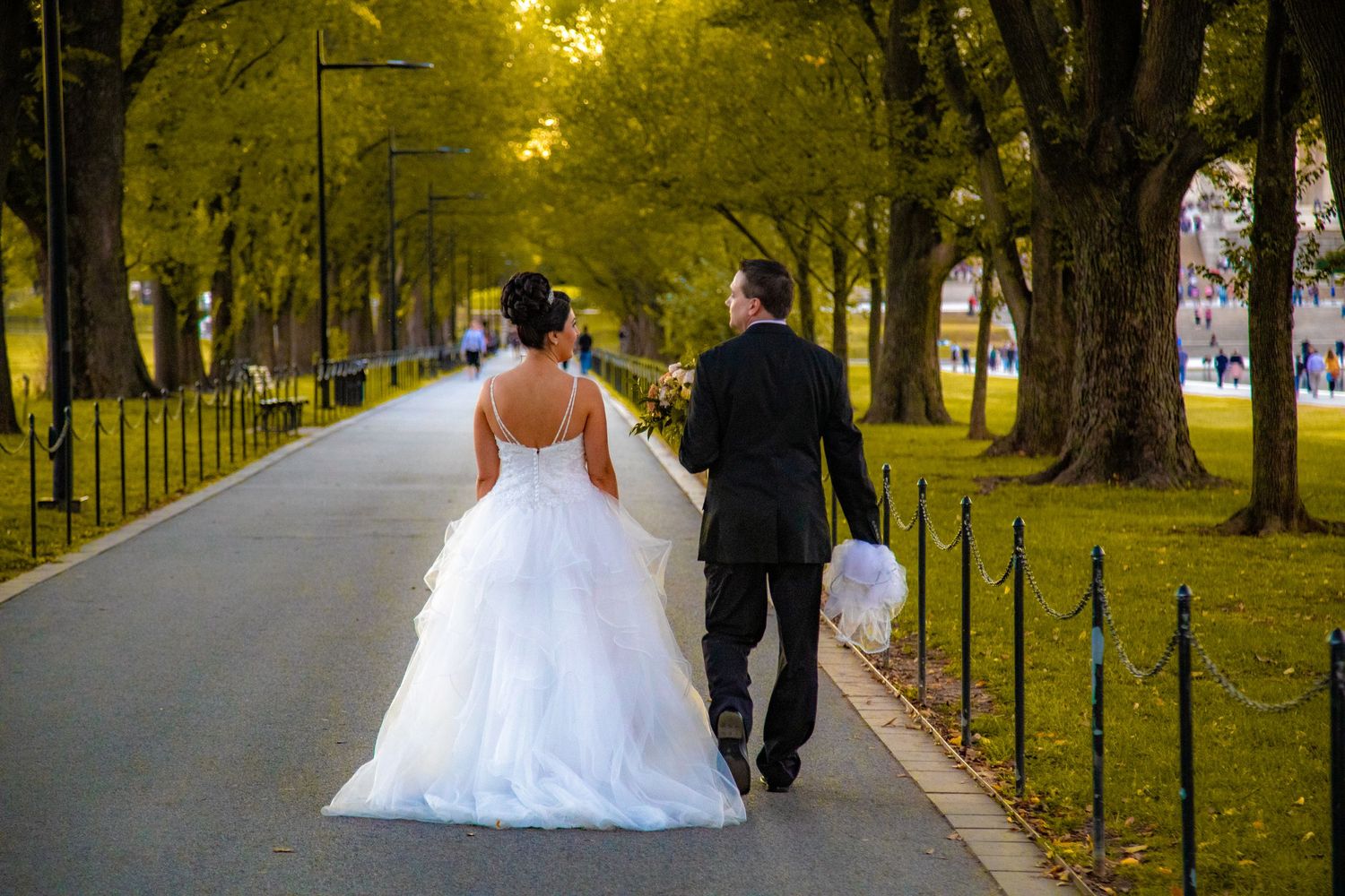 A couple walks down a tree-lined path on their wedding day with golden fall foliage overhead.