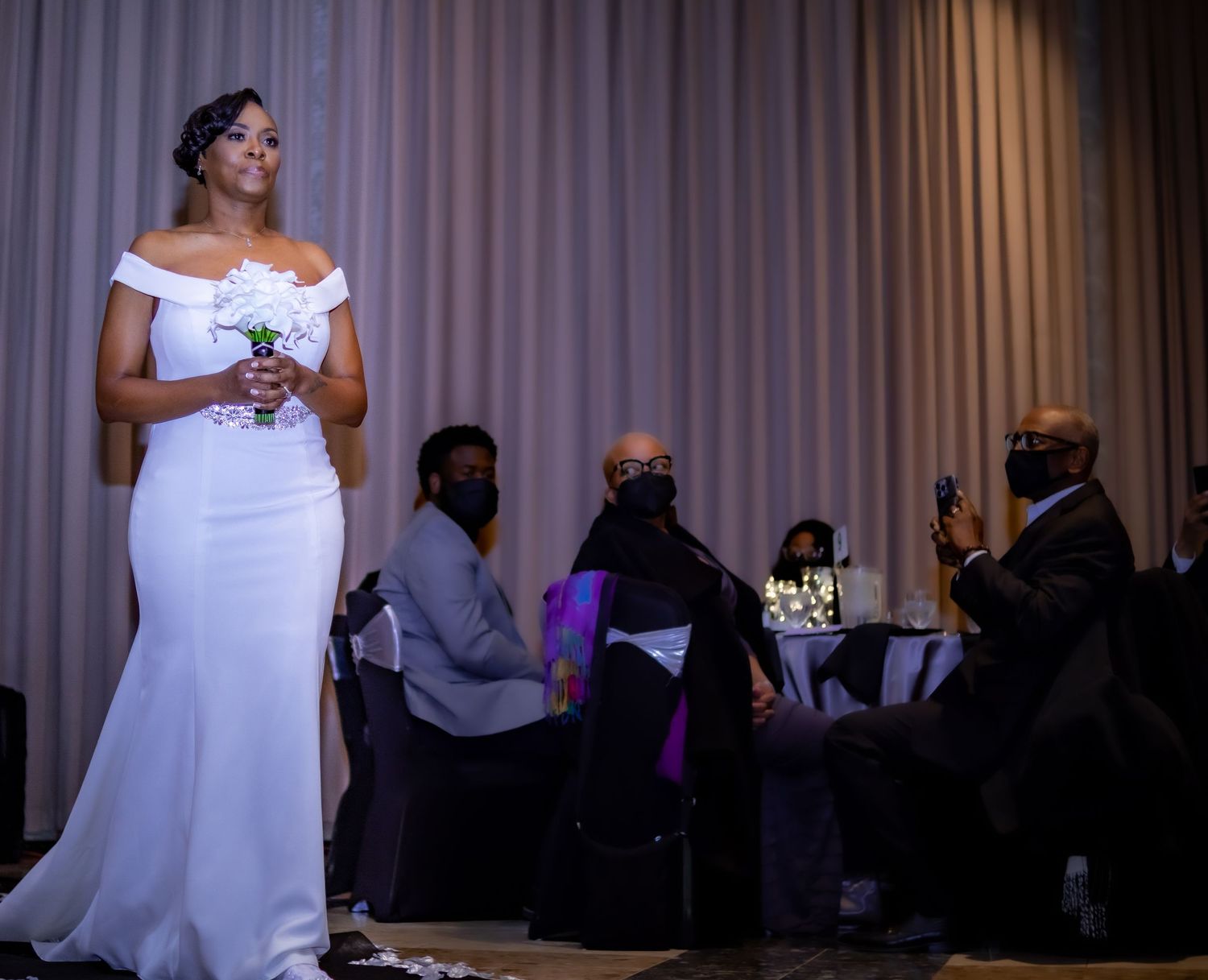 A bride in an off-shoulder white gown stands at a reception venue with guests seated in the background.