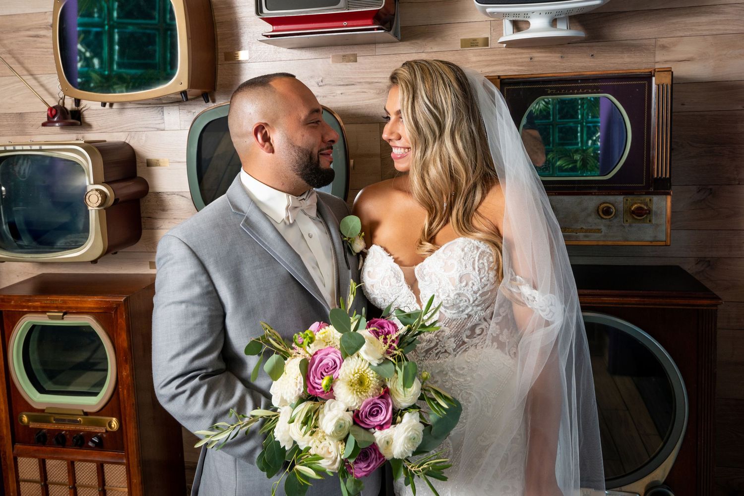 Newlyweds pose together in vintage-inspired setting with retro televisions and wood paneling.