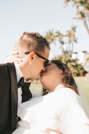 Romantic close-up of newlyweds sharing a kiss outdoors with palm trees visible.