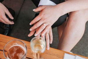 Close-up shot of hands resting on a wooden table with drinks, showing off a wedding ring.