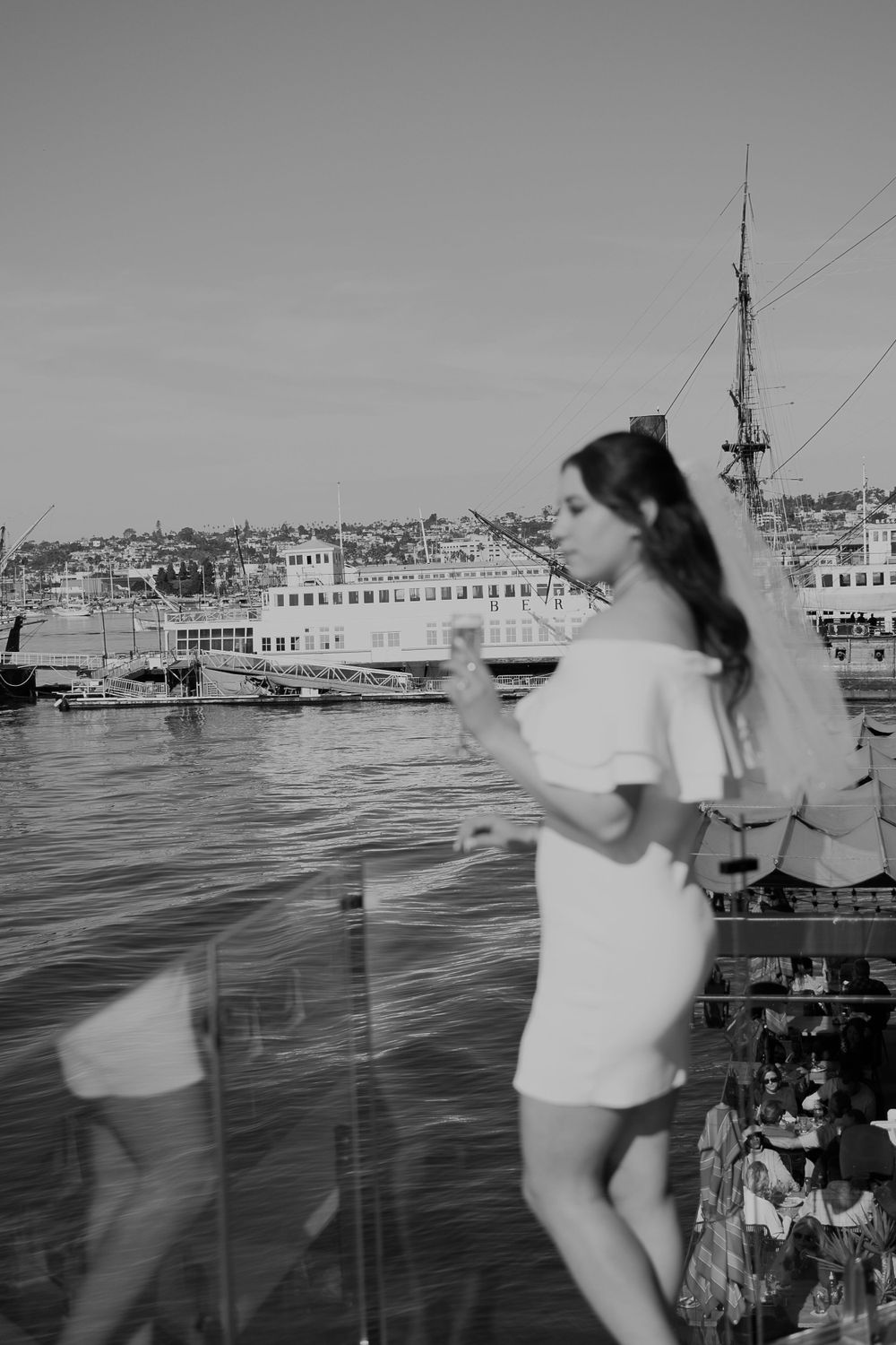Black and white photo of a woman in white dress standing by a harbor with boats in the background.