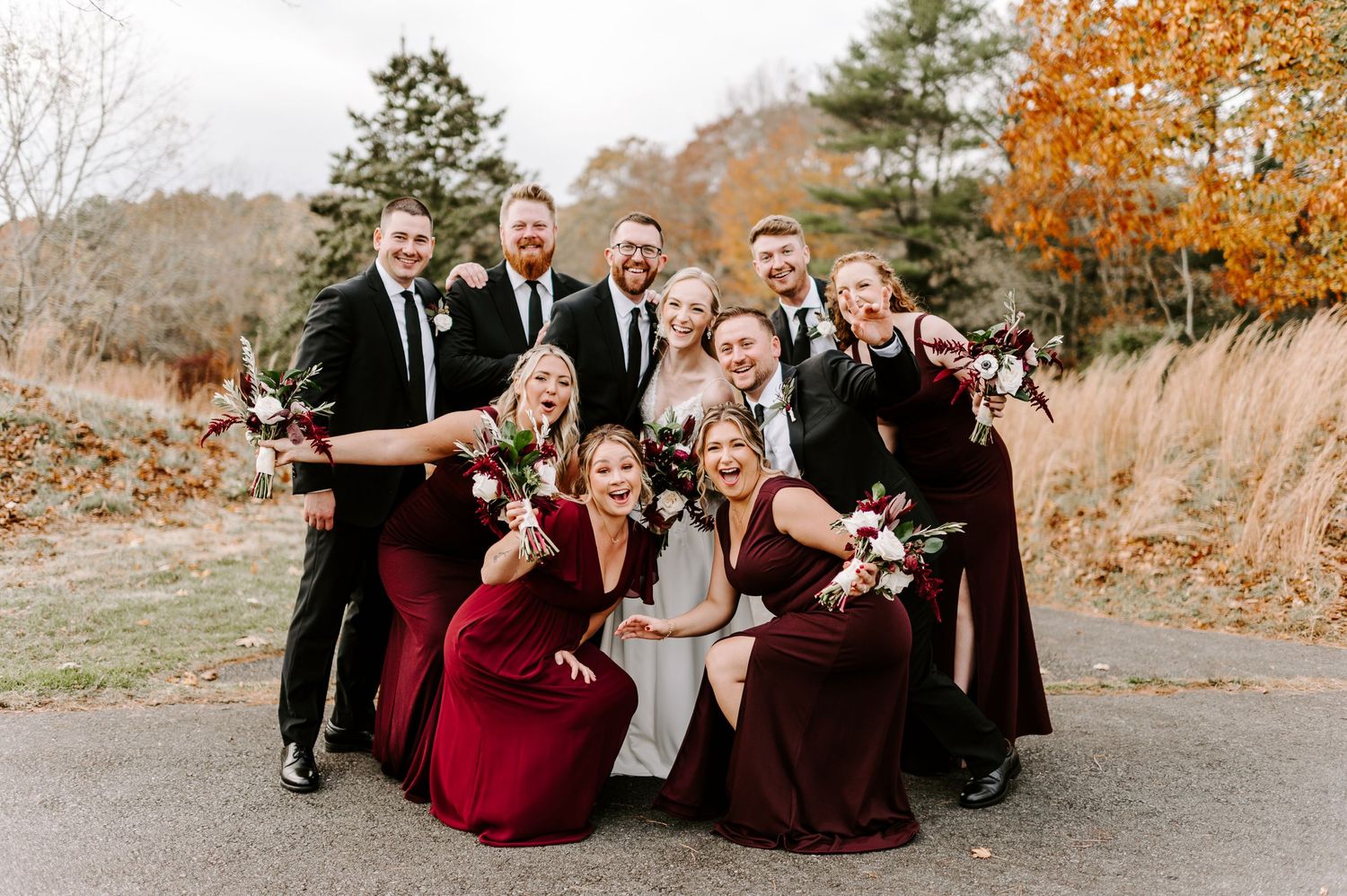 Wedding party poses playfully on path with bridesmaids in burgundy dresses and groomsmen in black suits.
