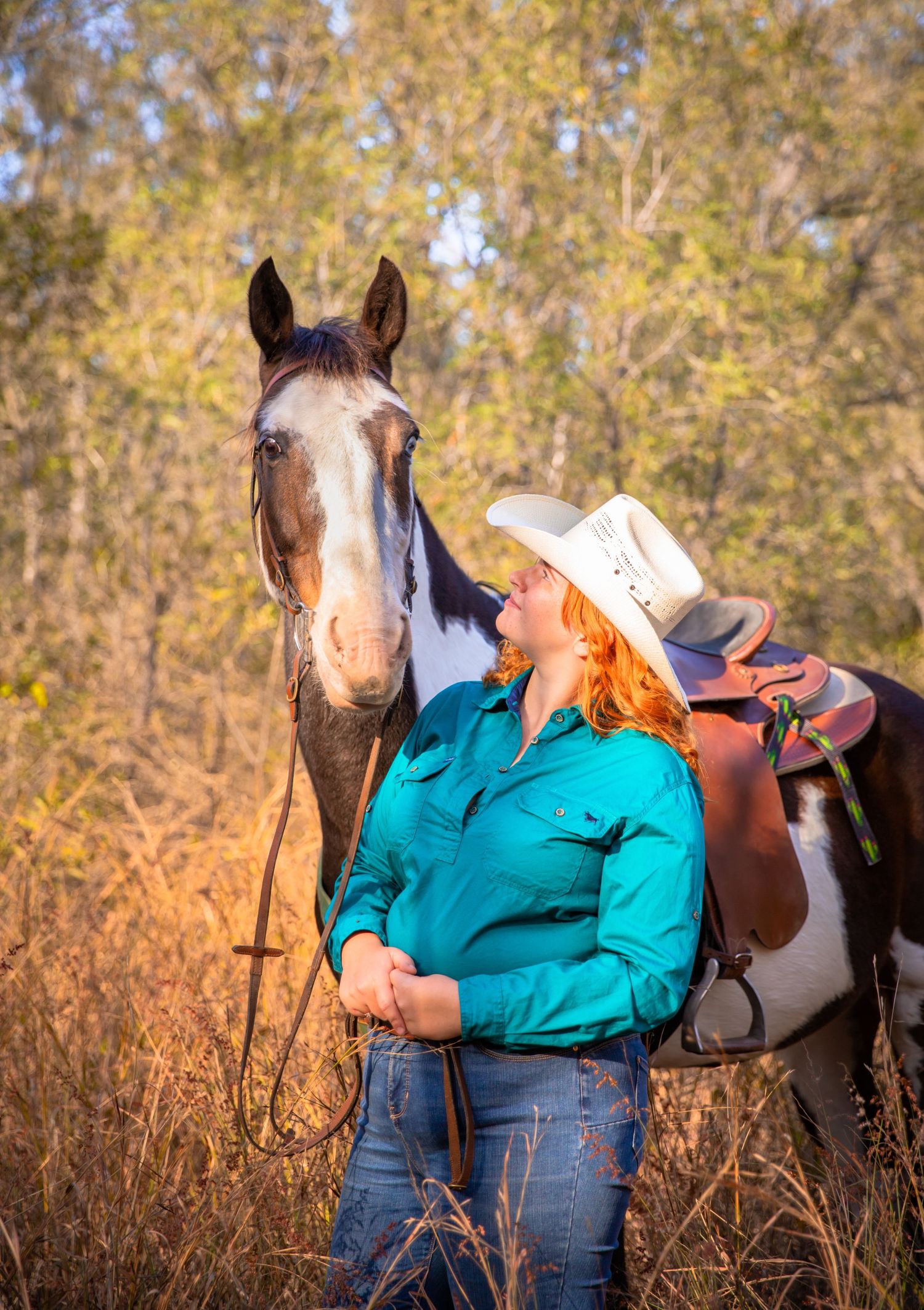 A girl in blue jeans and a blue shirt looks up at her paint horse who is facing towards the camera