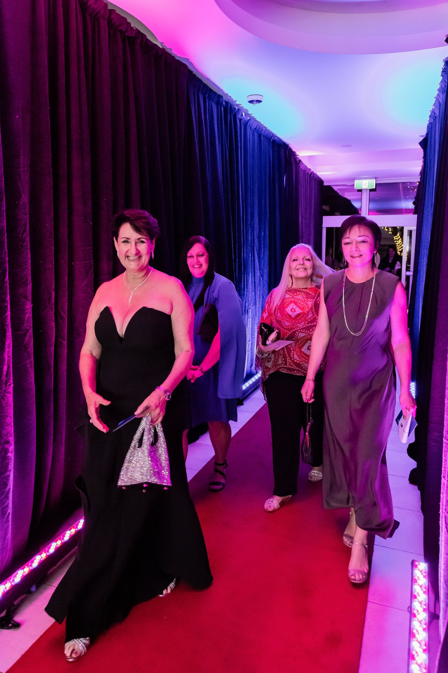 People walking down a red-carpeted hallway with blue curtains and pink lighting.