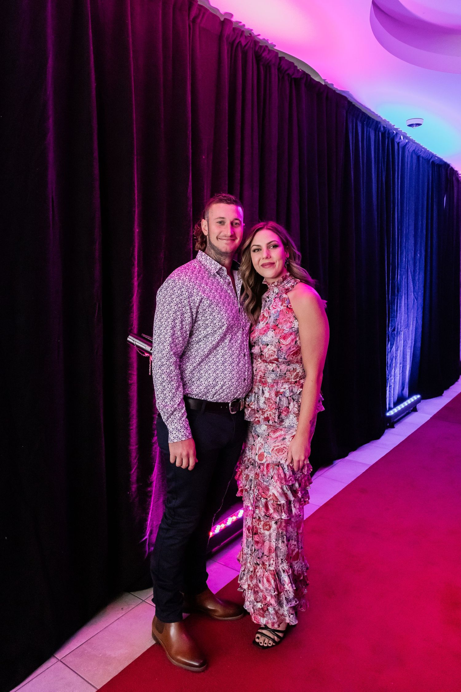 Couple posing on red carpet with dark curtain backdrop and pink ambient lighting.