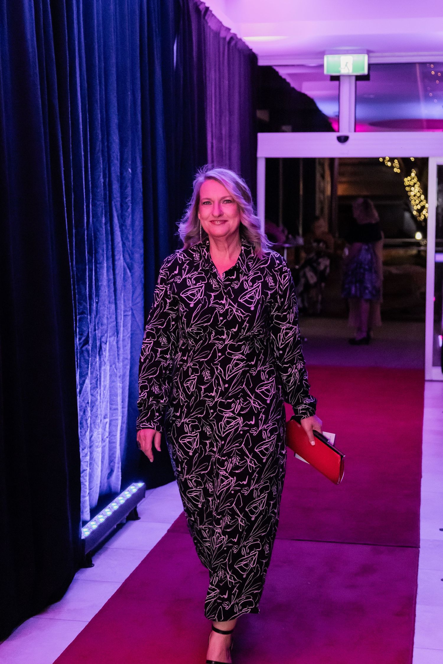 Person in patterned dress walking on red carpet with purple lighting and blue curtains.