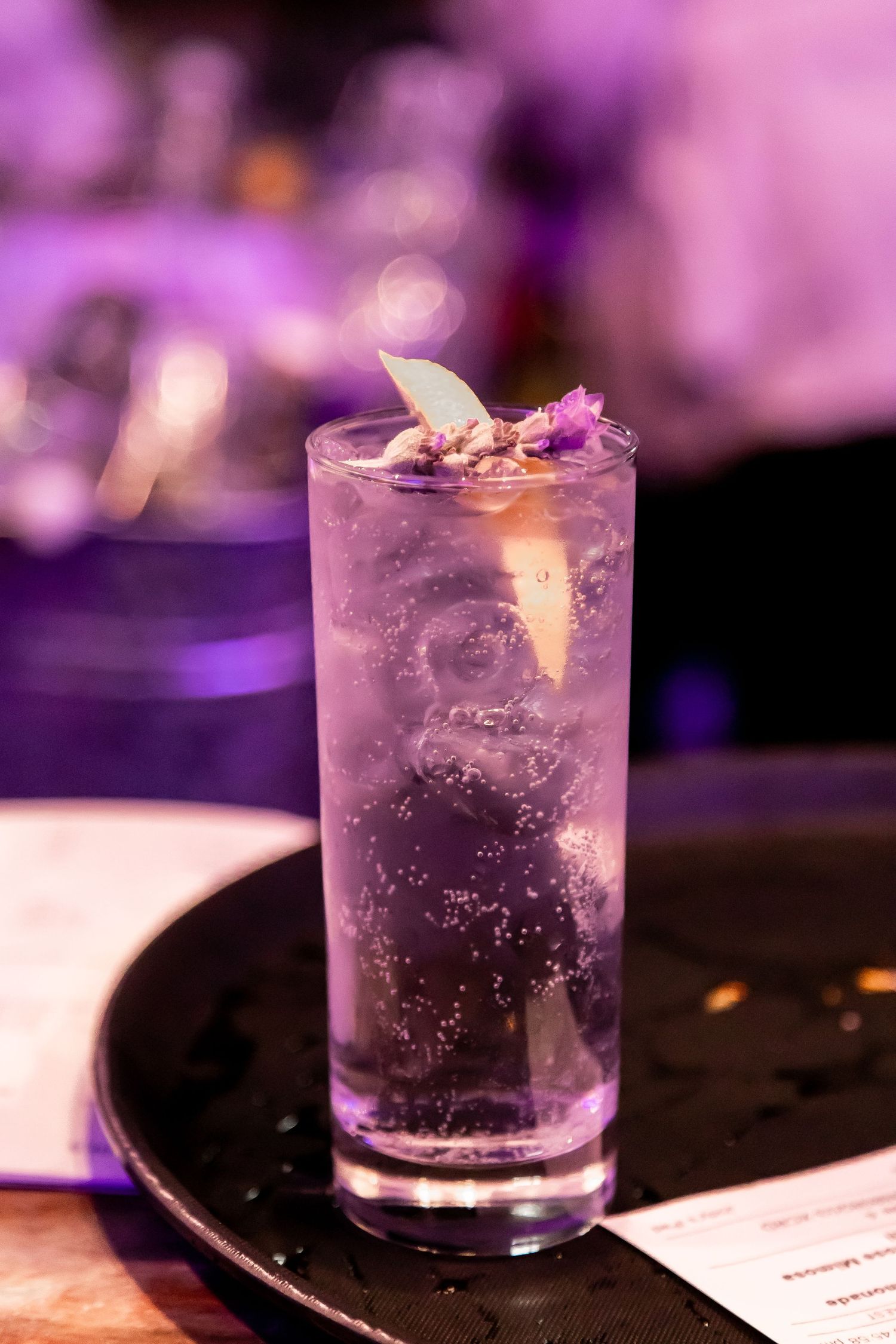 Purple-tinted cocktail with floral garnish served in tall glass on dark surface.