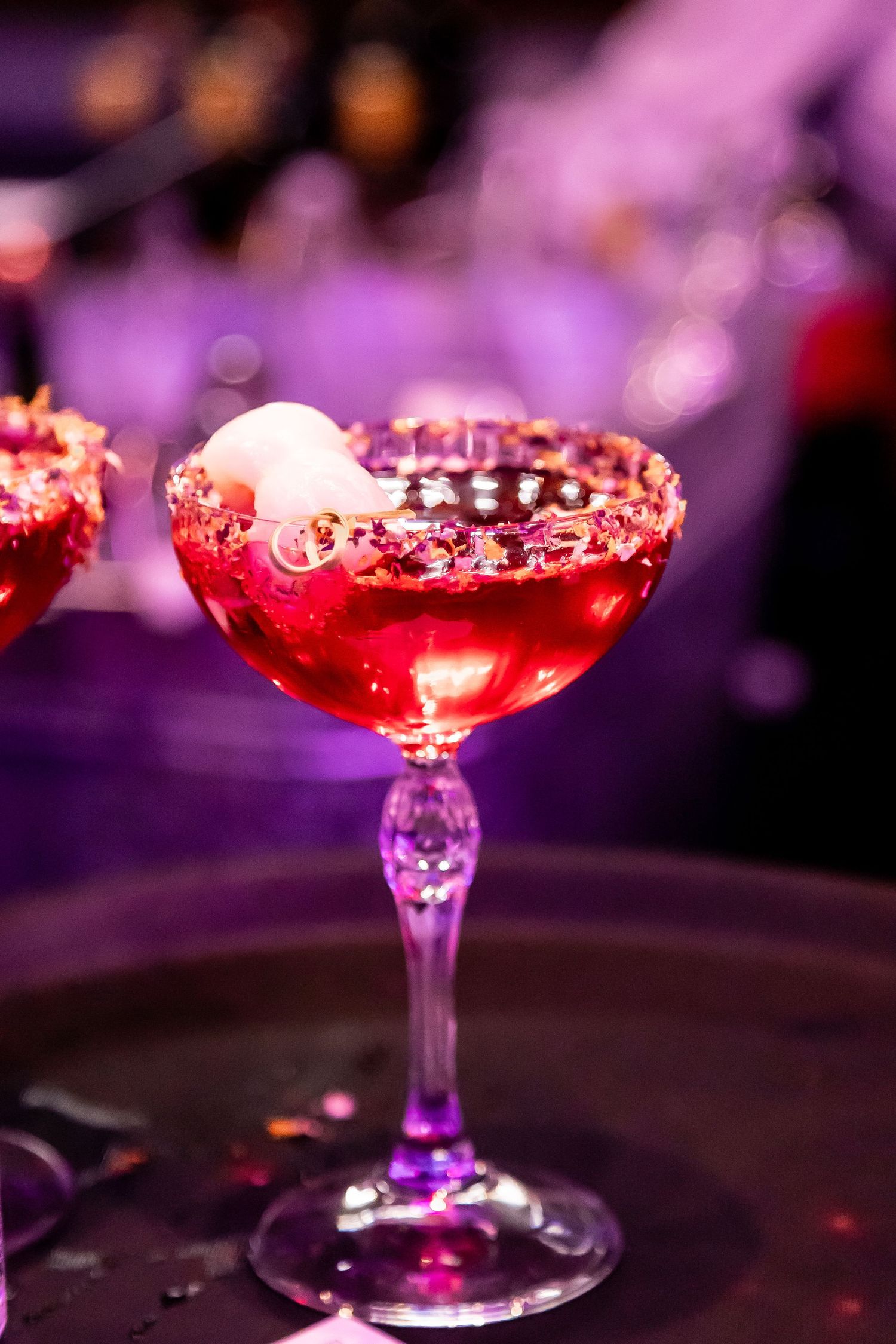Elegant red cocktail garnished with petals in coupe glass under purple lighting.