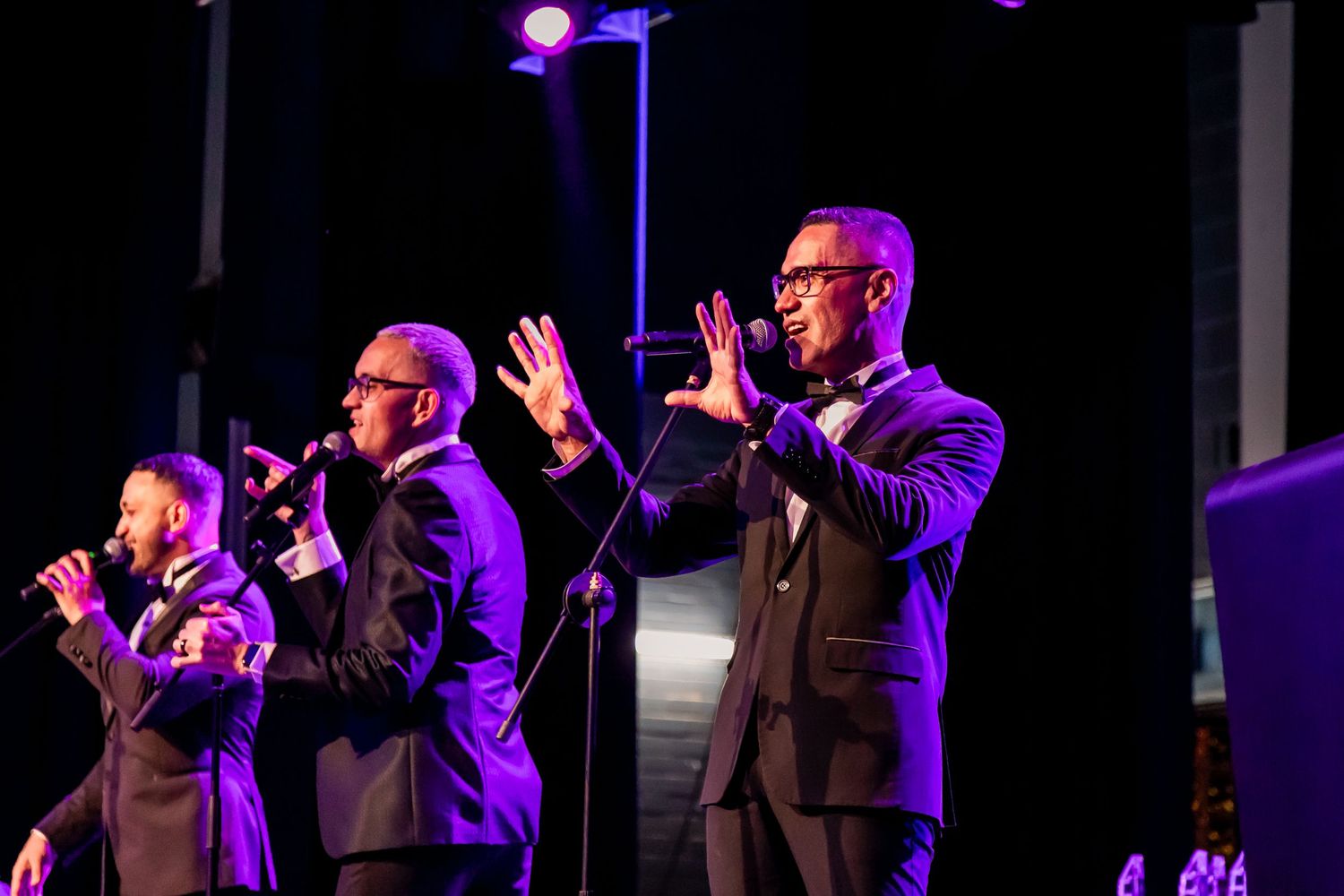 Three performers in suits singing on stage with purple lighting.