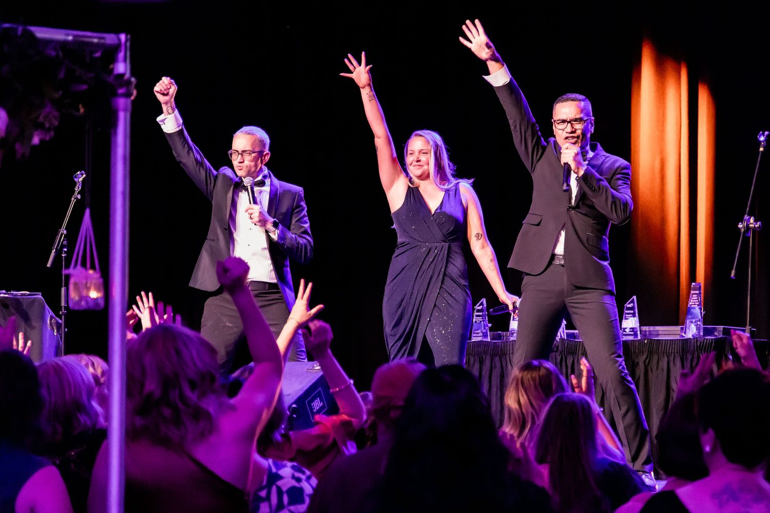 Energetic performers with raised arms on stage during live show with purple lighting.