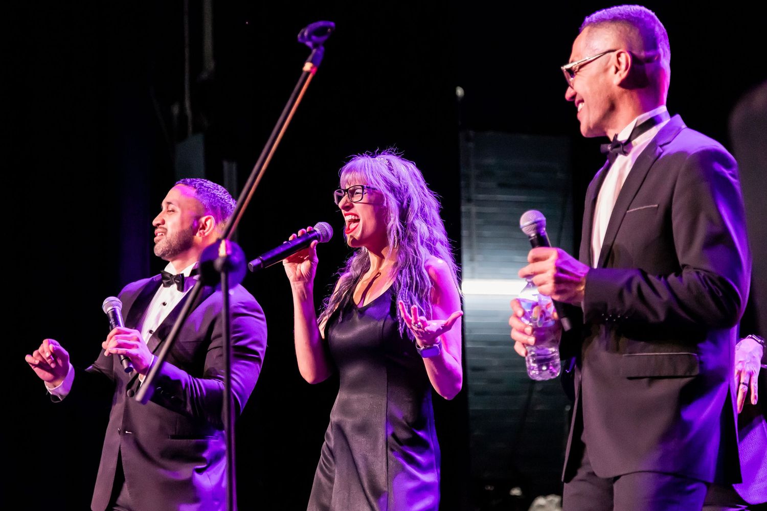 Musical performers in formal attire singing on stage with dramatic lighting.