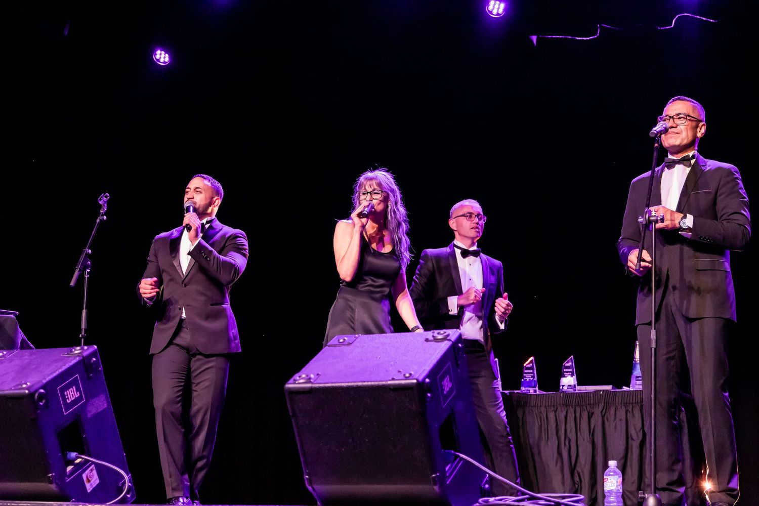 Performers in black formal attire singing on stage with microphones.