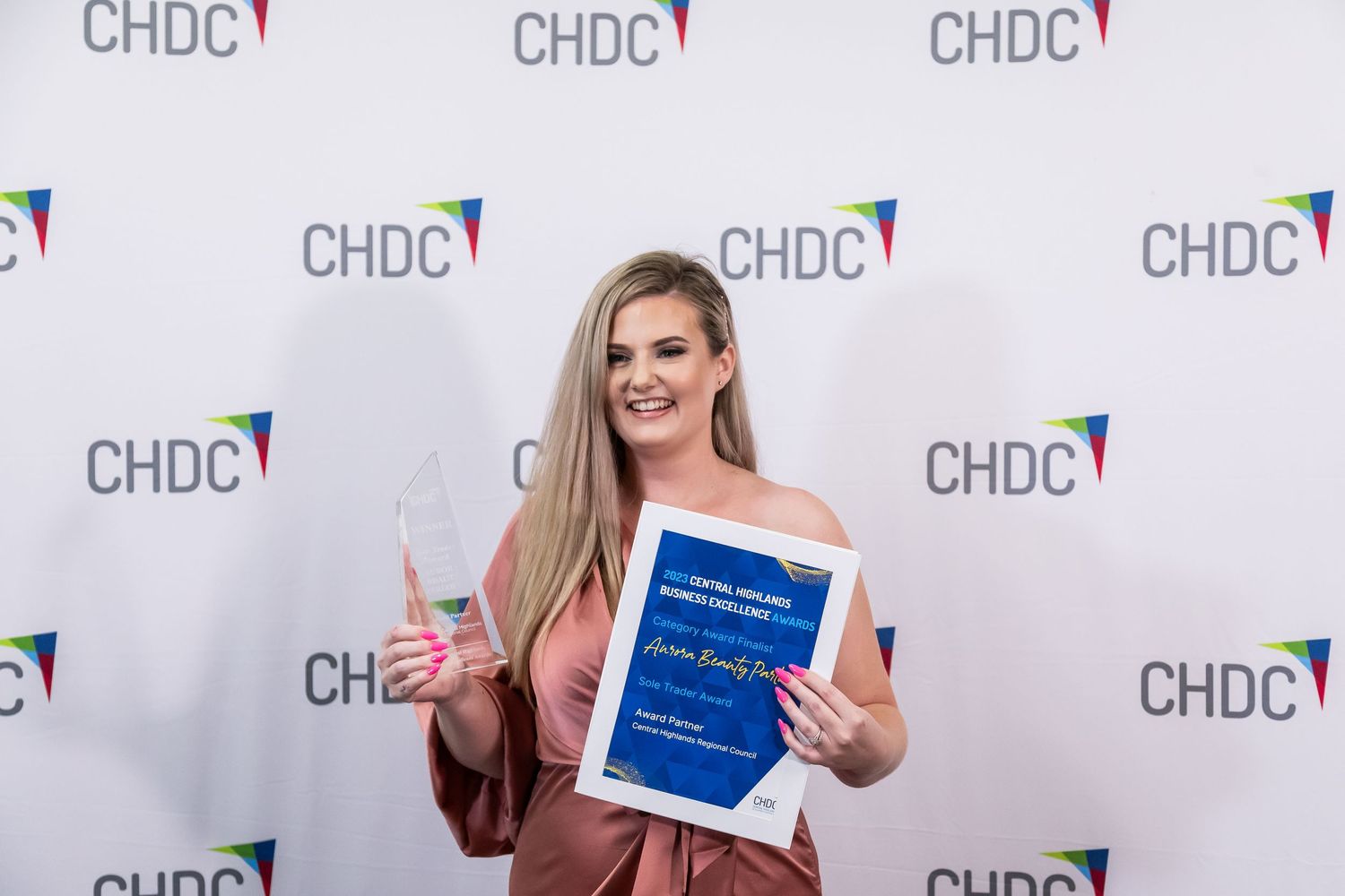 Award recipient in a brown dress holding up a blue certificate and peace sign at CHDC event.
