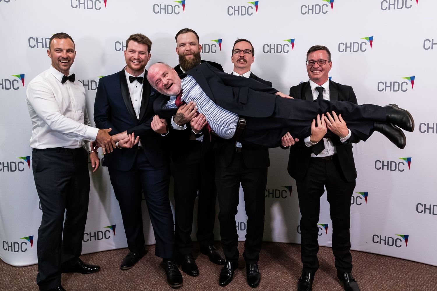 Five men in suits playfully carrying one person horizontally for a photo.