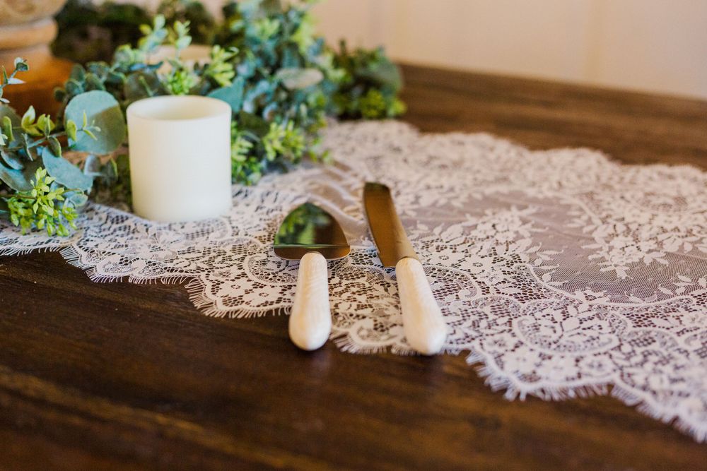 Wedding cake display with white candles and greenery on delicate lace tablecloth.