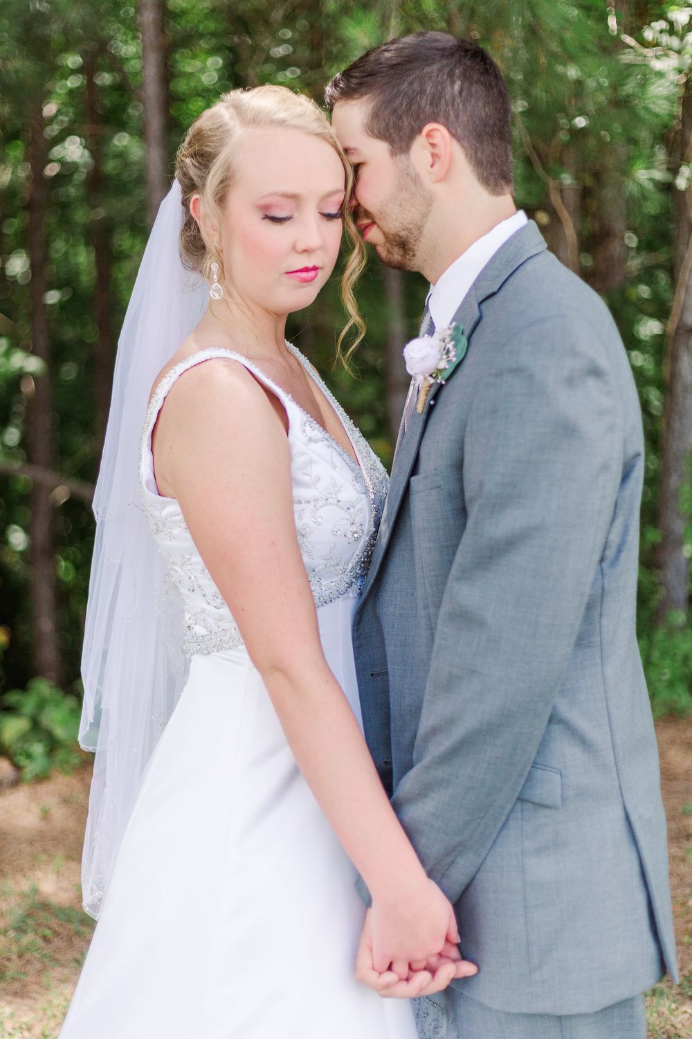 Romantic wedding couple embracing outdoors in grey suit and white lace dress among green foliage, by best San Antonio Photographer.
