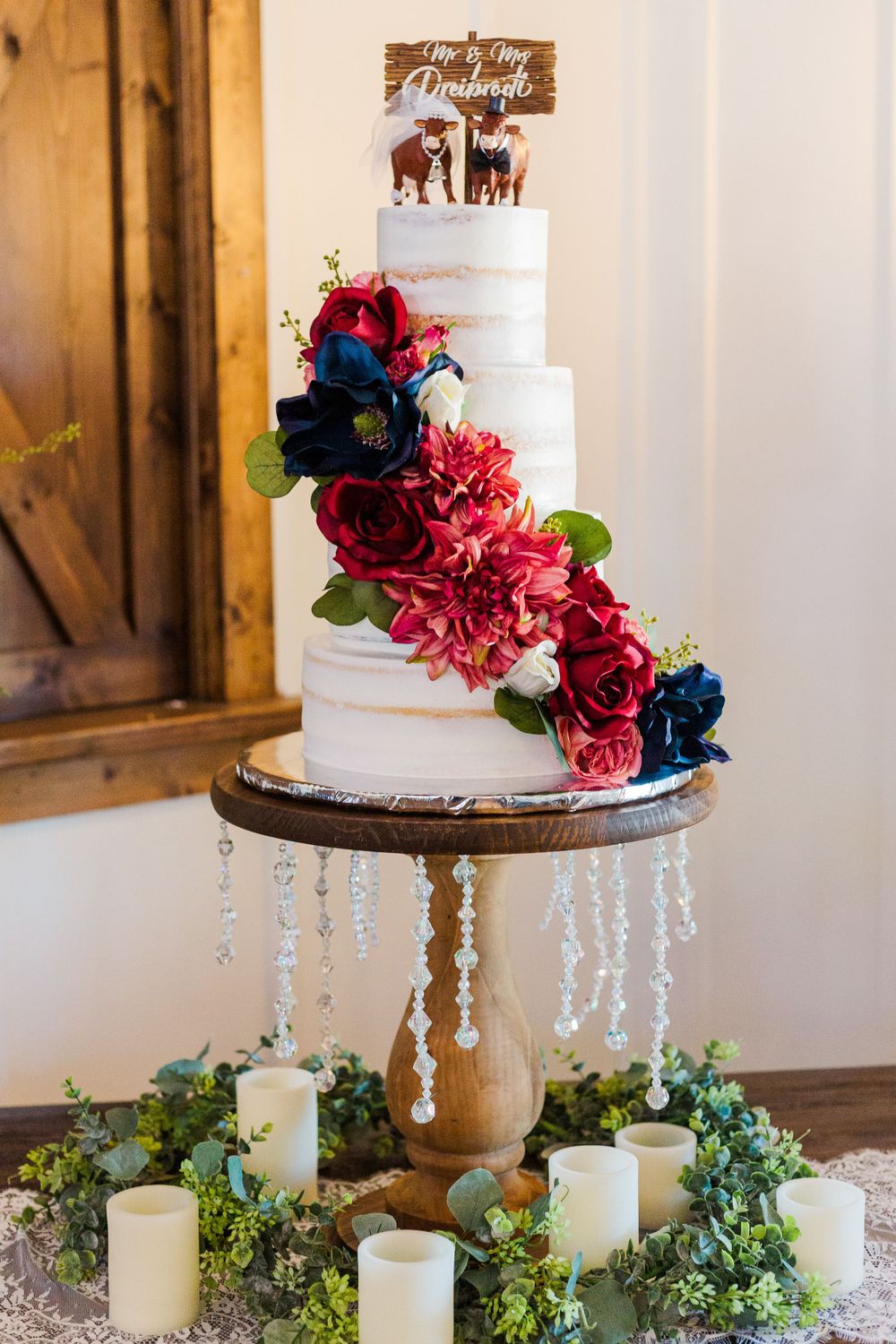 Elegant wedding cake decorated with deep red and navy blue flowers on a crystal-draped wooden stand.
