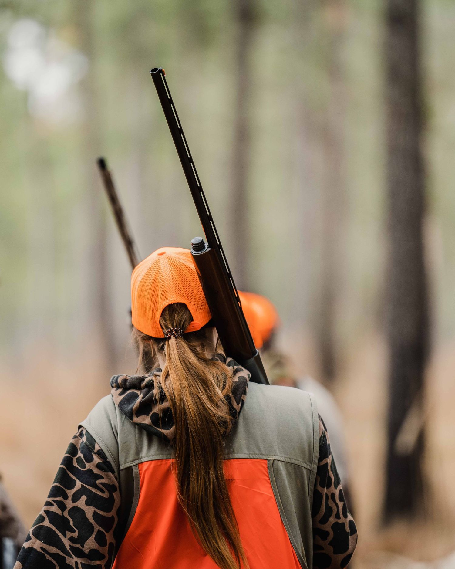 Hunter in orange safety vest and camouflage gear walking through forest with rifle.