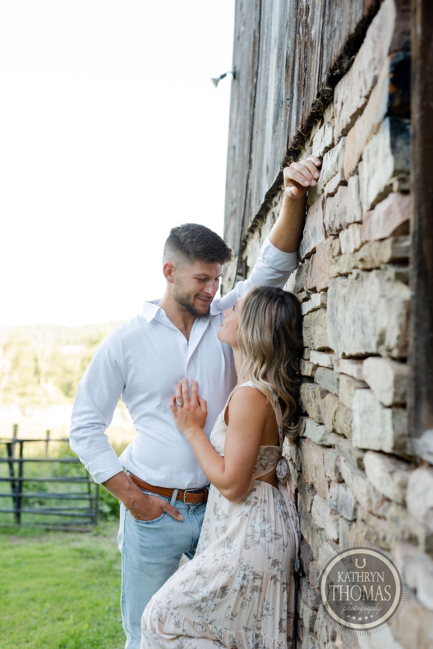 Romantic couple posing against rustic stone wall in countryside setting at golden hour.