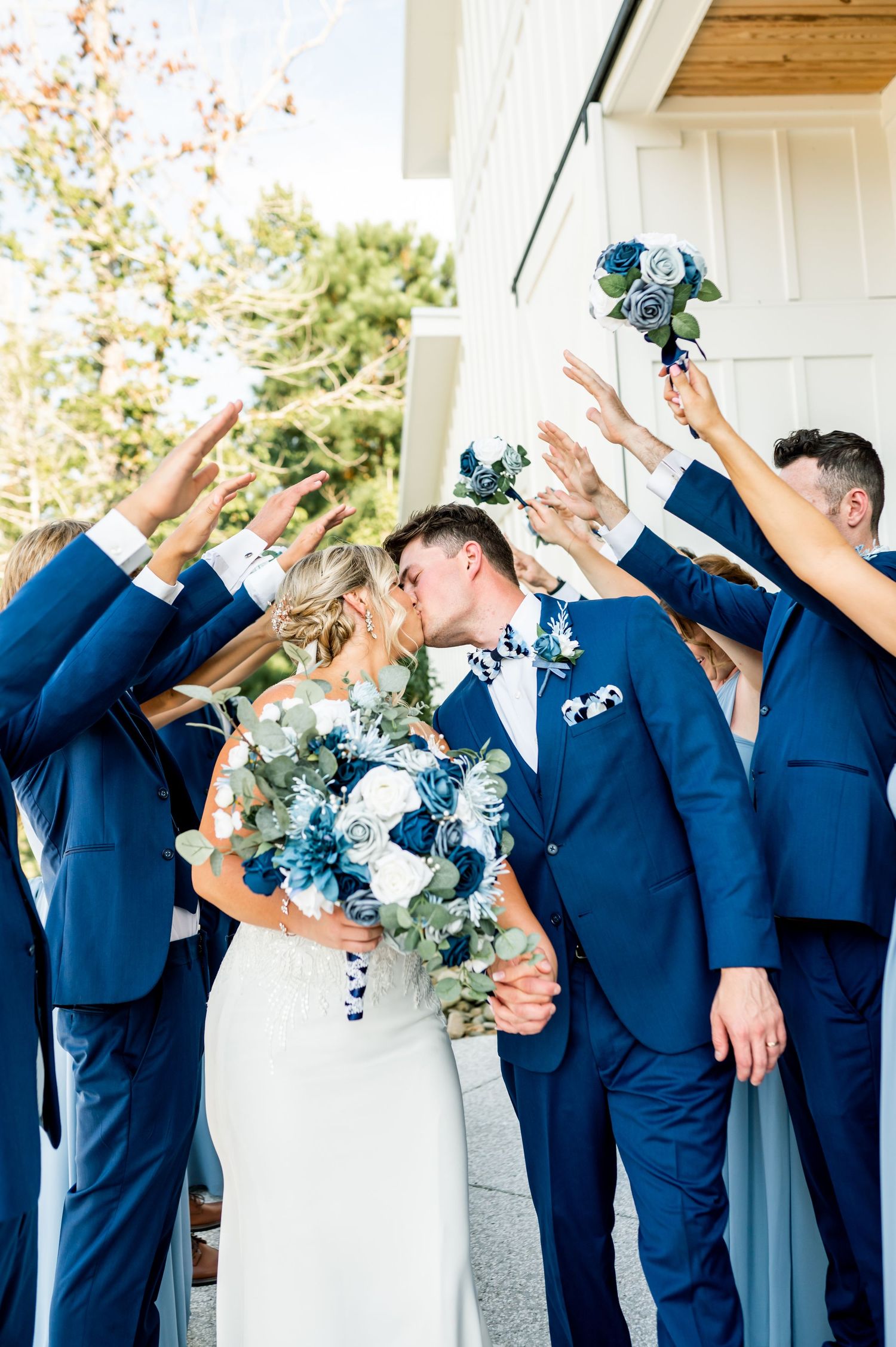 Bridal party in blue attire strikes dynamic poses against white building exterior.