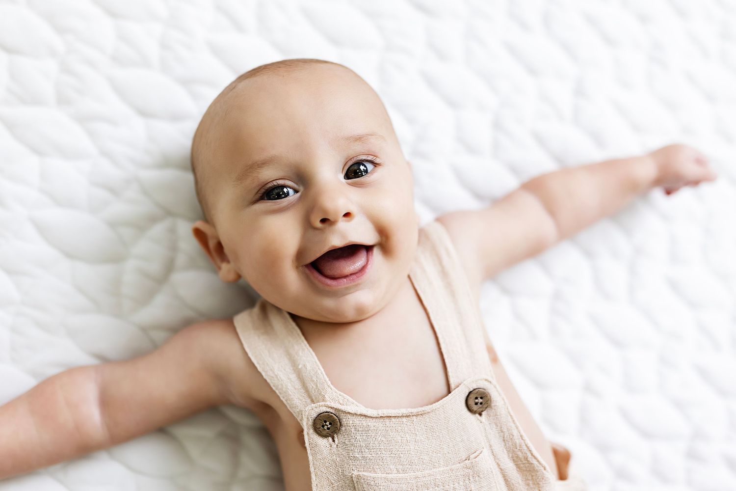 Smiling baby in beige overalls lying on white textured background.