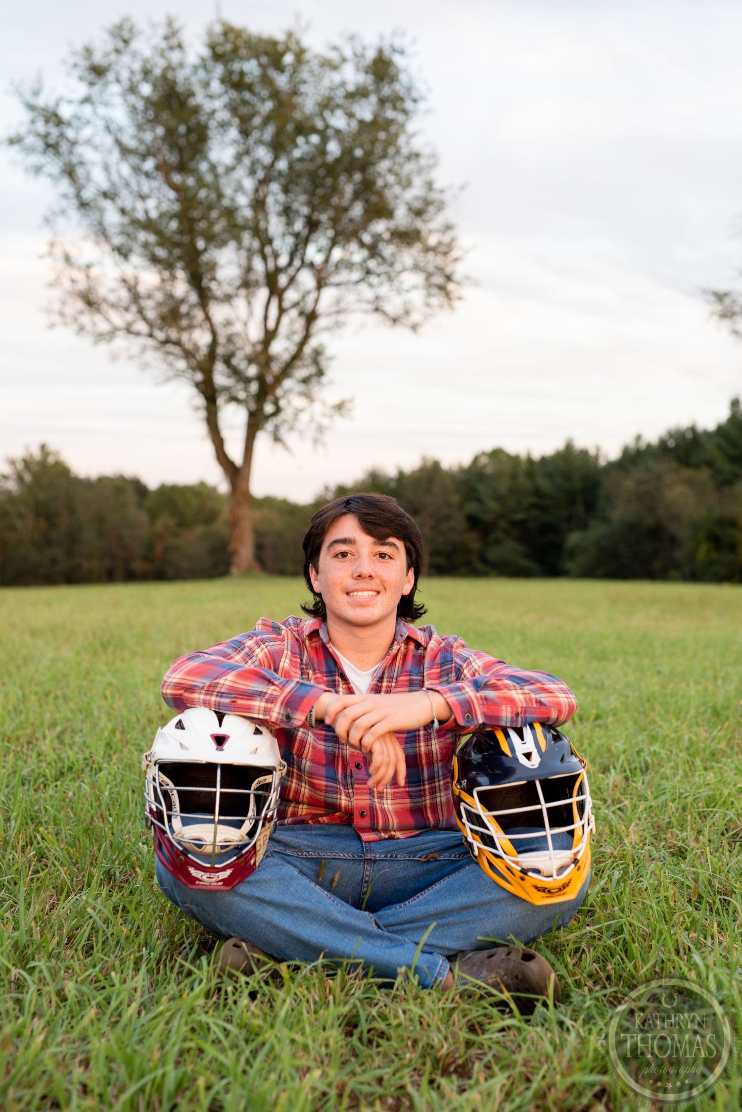 Person in plaid shirt sits cross-legged holding two sports helmets in grassy field.