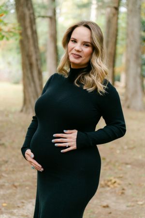 Pregnant woman in a black ribbed dress gently holding her baby bump in a park.