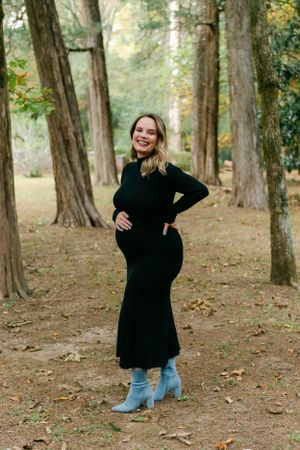 Smiling pregnant woman in a black maternity dress posing in a wooded outdoor setting.