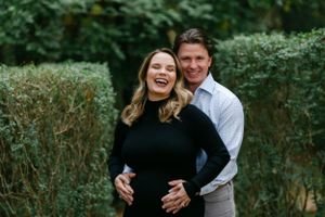 Laughing pregnant couple embracing outdoors surrounded by tall green hedges in a garden.