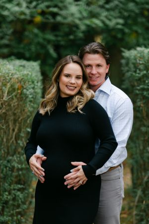 Smiling couple posing together with hands on pregnant belly in a lush garden setting.