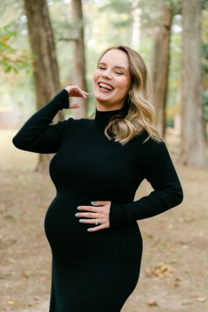 Laughing pregnant woman in a black turtleneck dress cradling her baby bump outdoors.