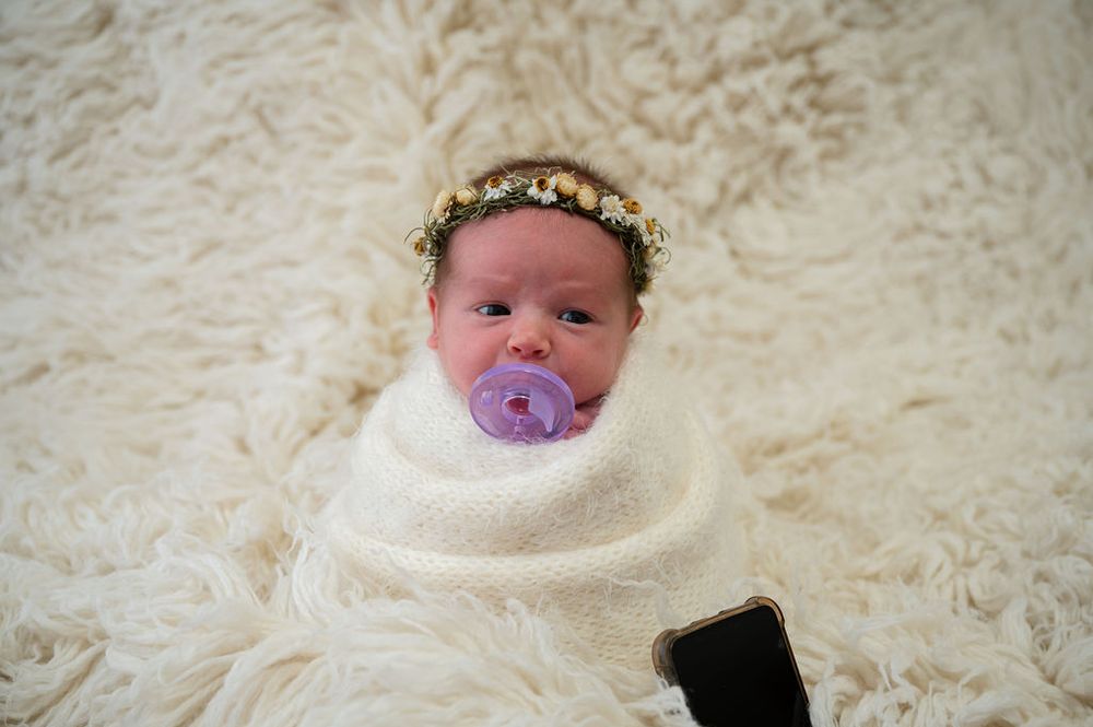 A newborn wrapped in white with flower crown and pacifier rests on textured cream fabric.