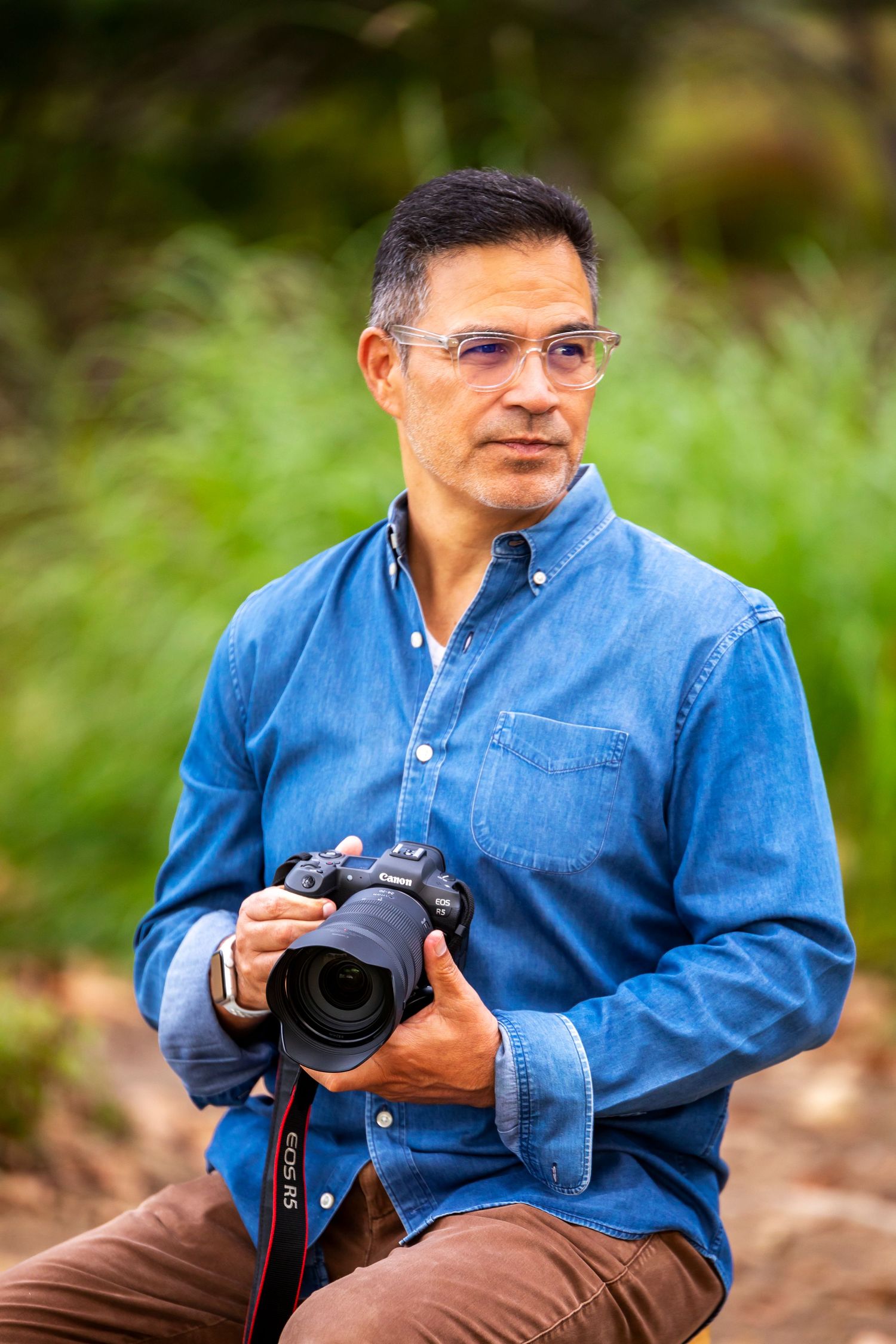 Man in blue denim shirt holding a Canon DSLR camera outdoors with green foliage background.