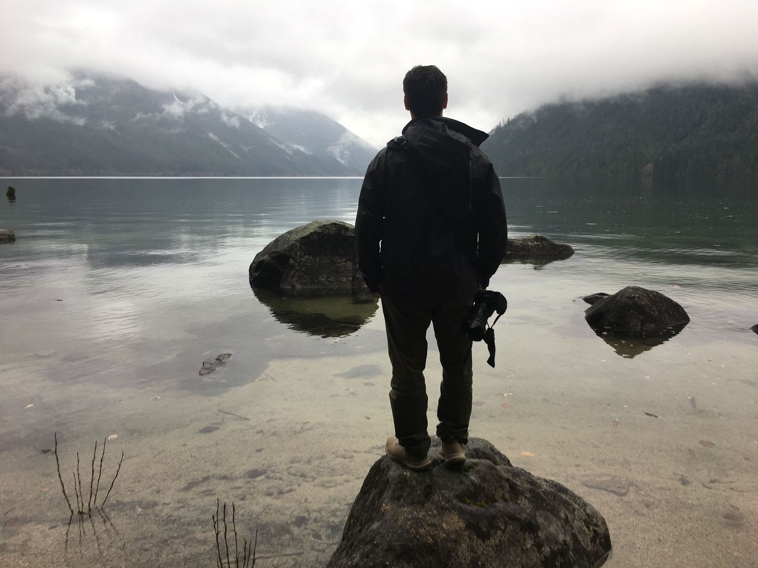 Myself (a man) looking out to the mountains on a grey day standing on a rock along the water.
