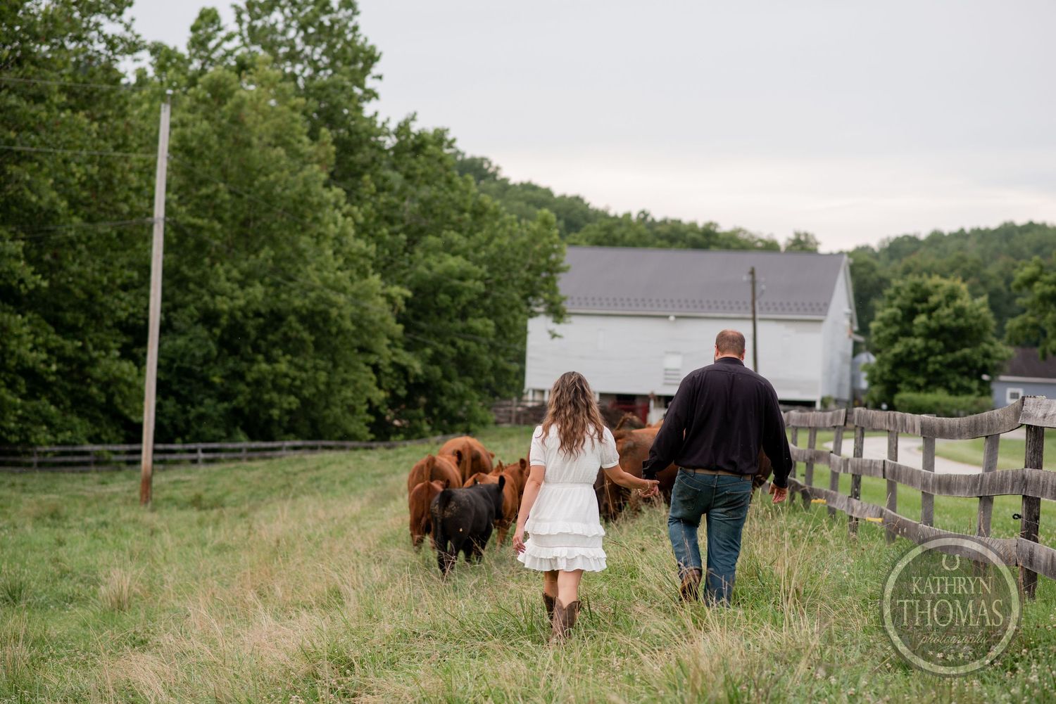 A couple walks with their dog towards a white barn on a grassy field.