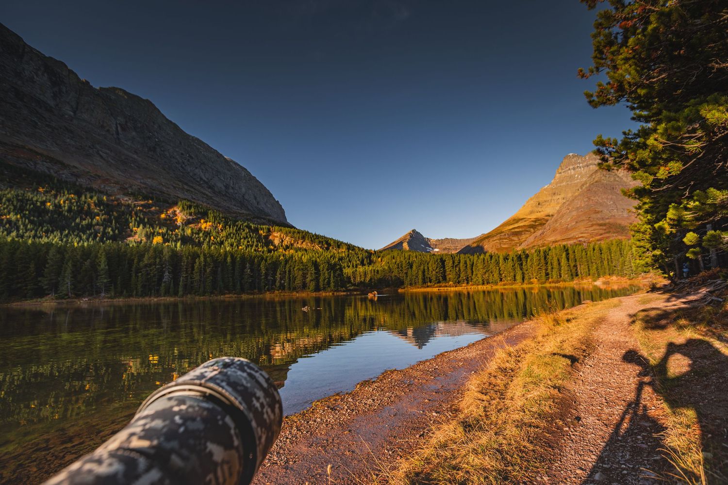 Golden sunlight illuminates a trail along an alpine lake surrounded by mountains and evergreen forest.