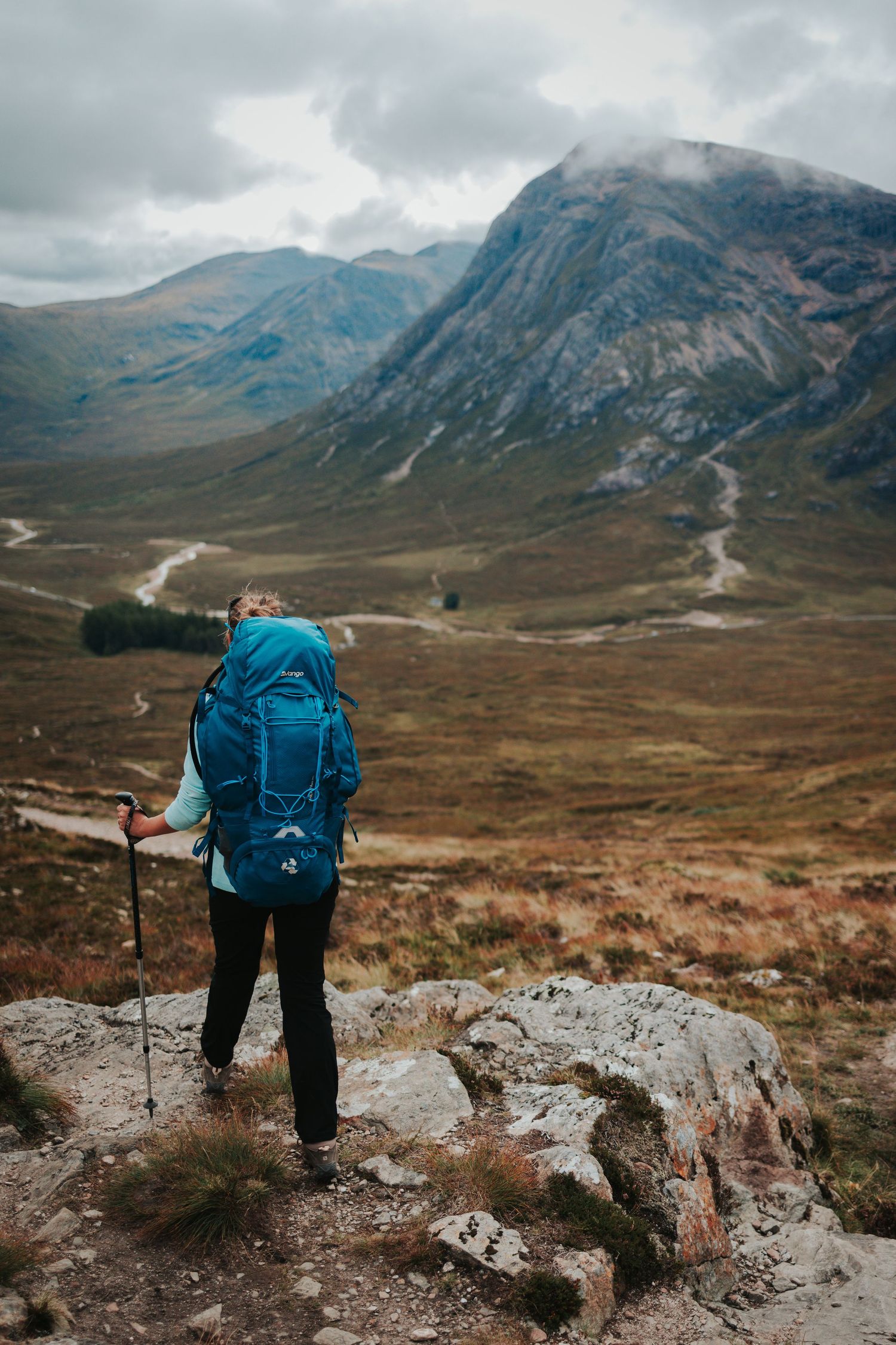Hiker with blue backpack stands on rocky outcrop overlooking dramatic mountain valley landscape.