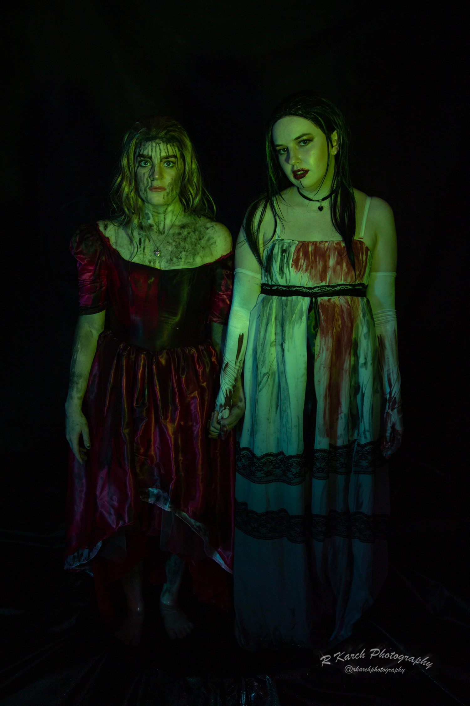 Gothic styled photograph of two figures in blood-stained period dresses under green light.